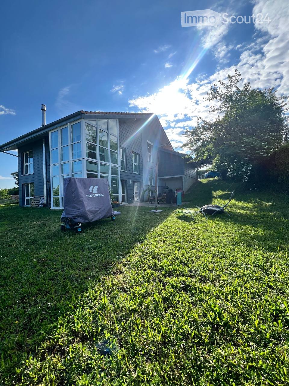 A modern house with large windows and a covered patio area, surrounded by a well-maintained lawn and outdoor furniture.