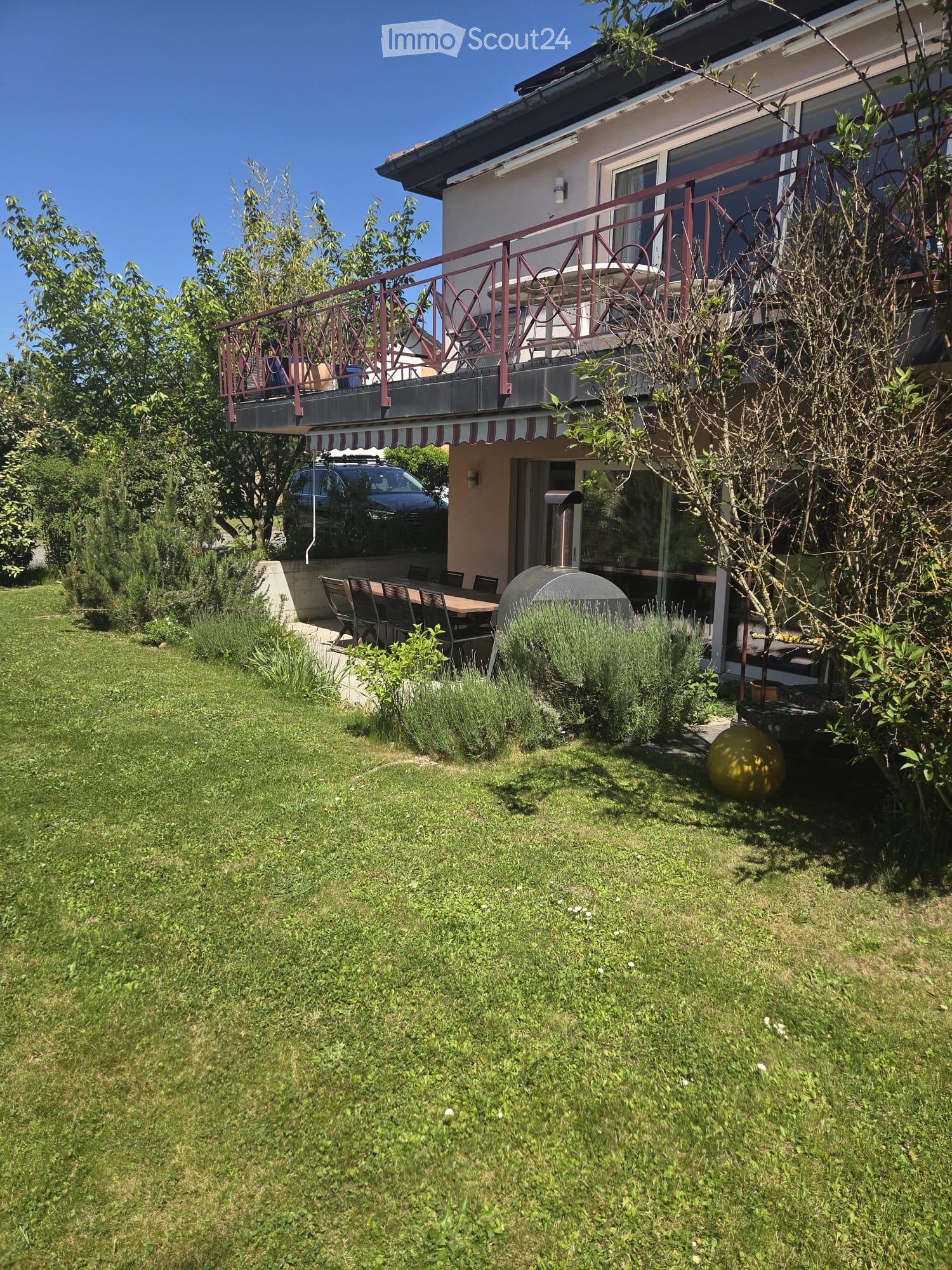 Lush garden with a well-maintained lawn, a patio area with a table and chairs, a round outdoor ball, and a balcony with a railing and outdoor furniture.