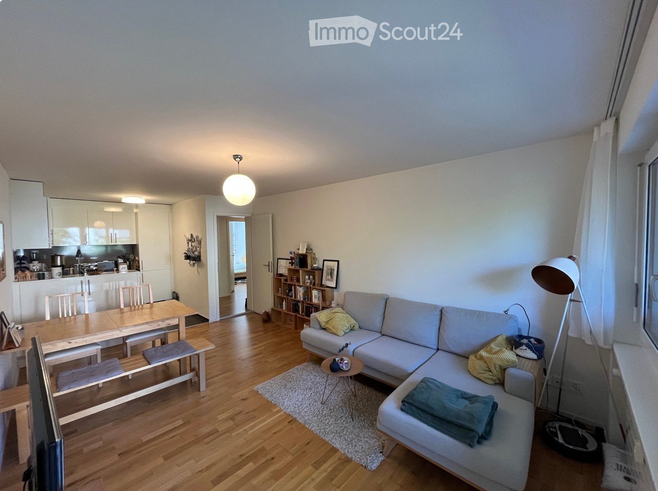 Bright, open-plan living and dining area with wooden flooring, a grey sectional sofa, a small coffee table, a dining table with chairs, a bookshelf, and a modern kitchen area with cabinets and appliances.