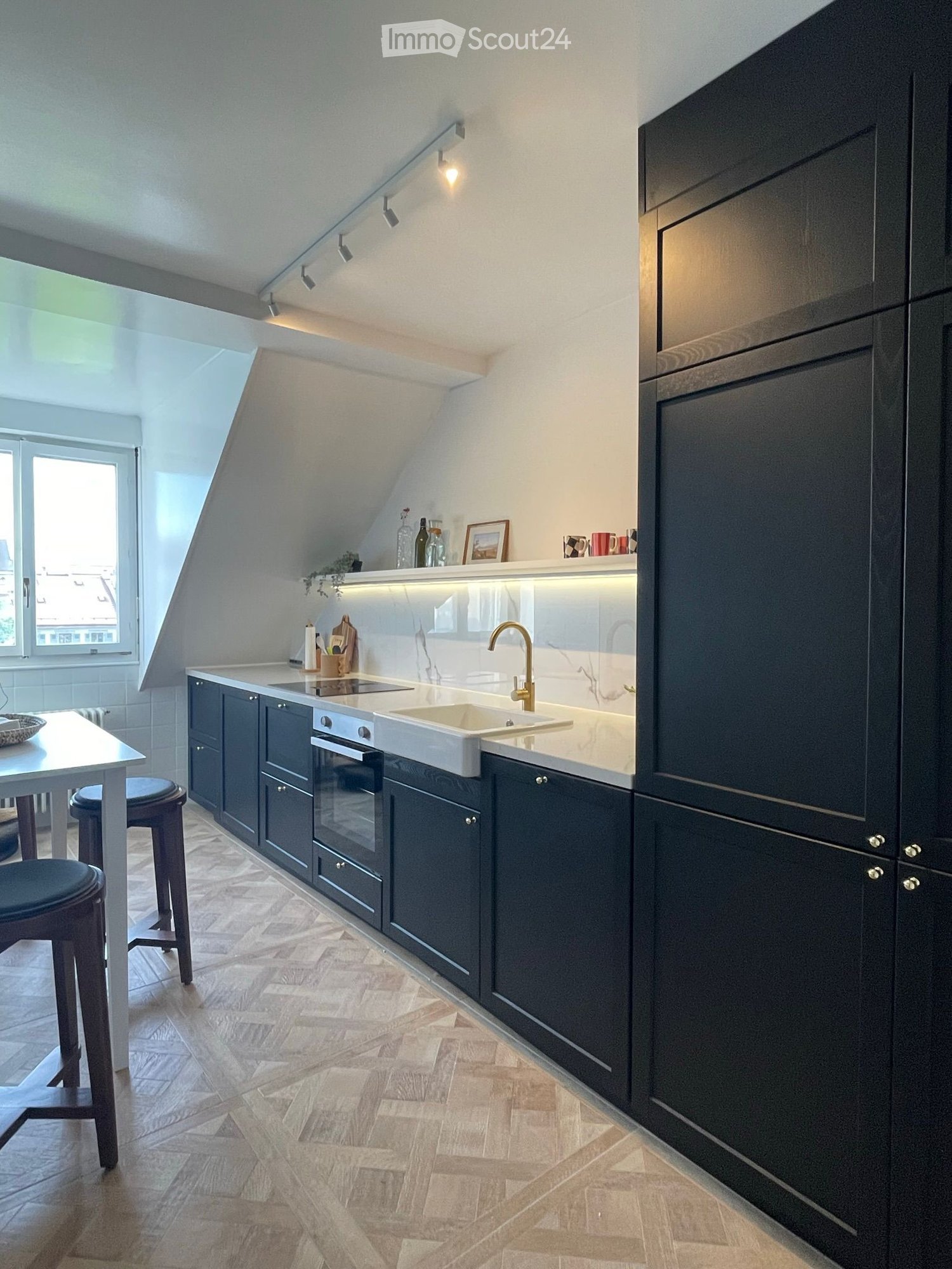 Modern kitchen with dark cabinets, white countertops, and a built-in sink and oven. Features include a sleek faucet, a small dining area with a table and stools, and decorative items on the countertop.