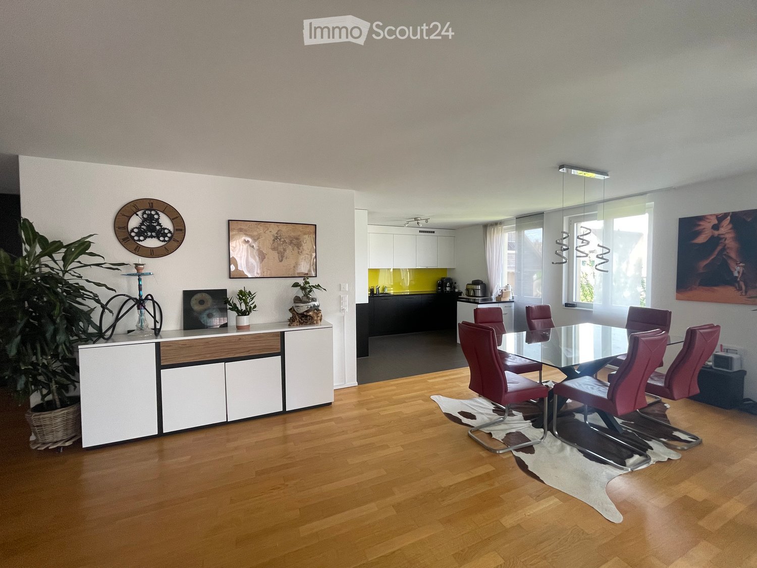 Open-plan living and dining area with wooden flooring, white and wood cabinetry, a glass dining table with red chairs, and a kitchen area with white cabinets and a yellow backsplash.