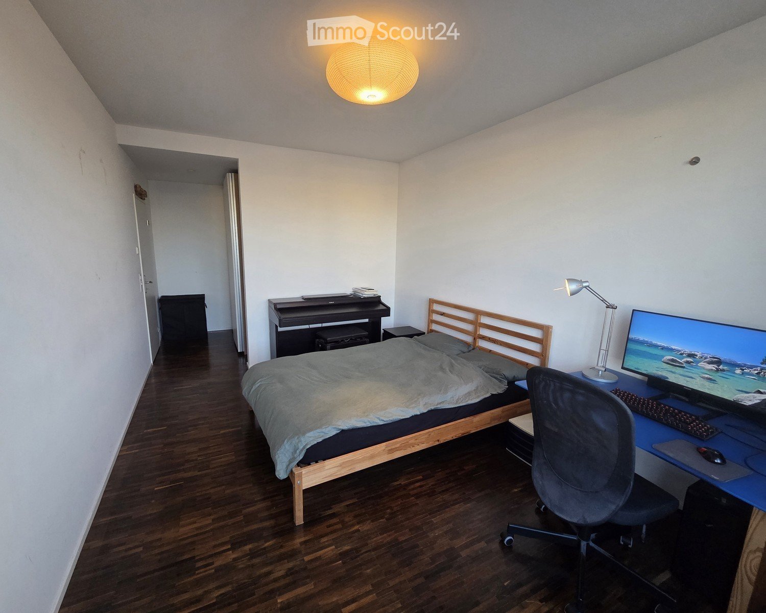 A modern bedroom with a wooden bed, desk setup, and a small piano. The room features wooden flooring, white walls, and a hanging light fixture.