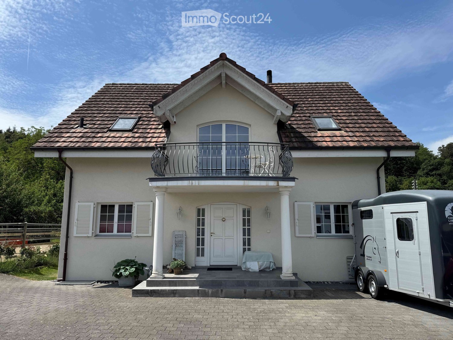 Two-story house with a tiled roof, white exterior, balcony, and covered entrance. A trailer is parked nearby.