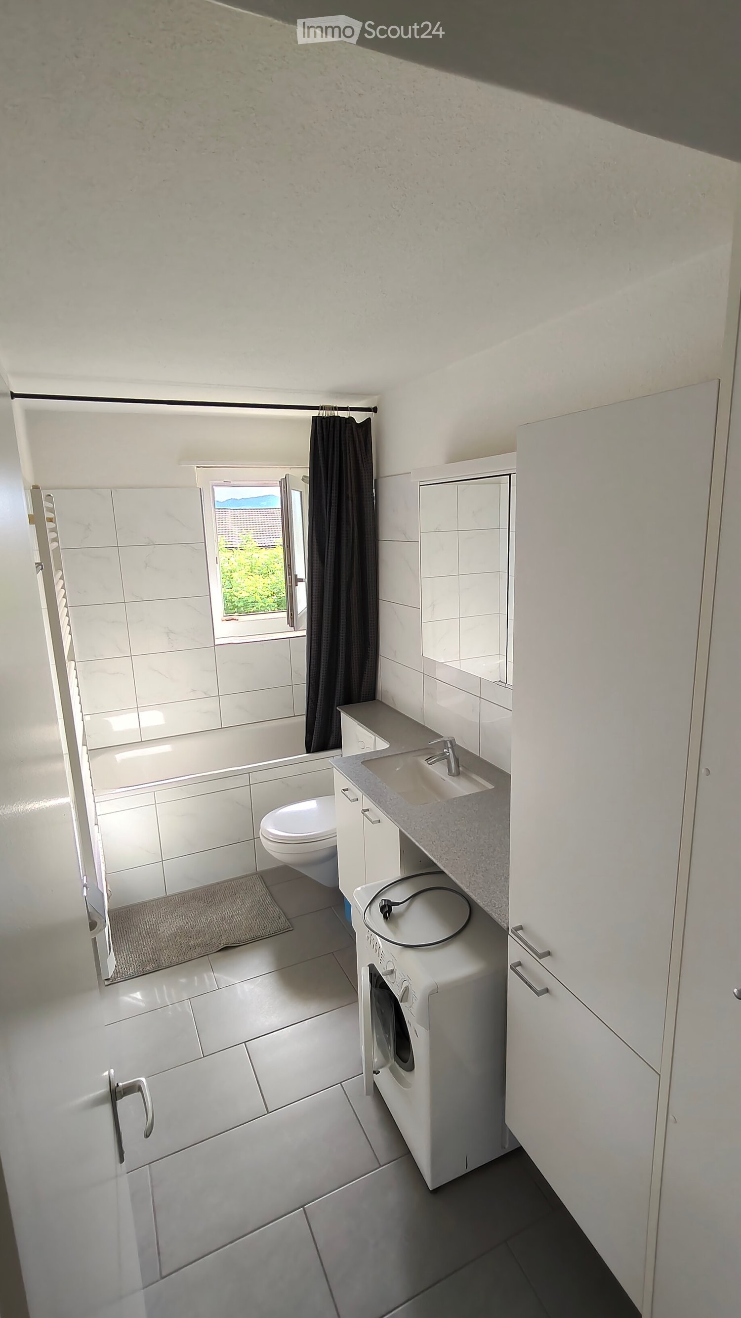 Modern bathroom with white tiles, a sink, toilet, bathtub, washing machine, and a window with a curtain.
