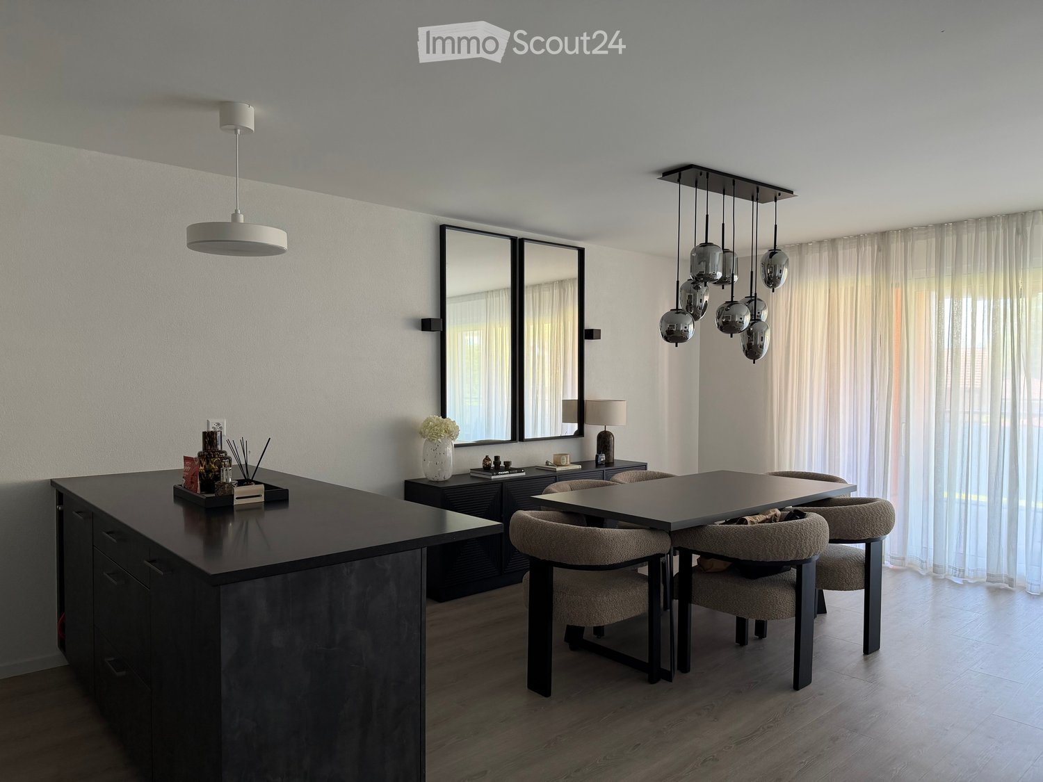 Modern dining area with a sleek black dining table, four chairs, a large kitchen island, and contemporary lighting fixtures.