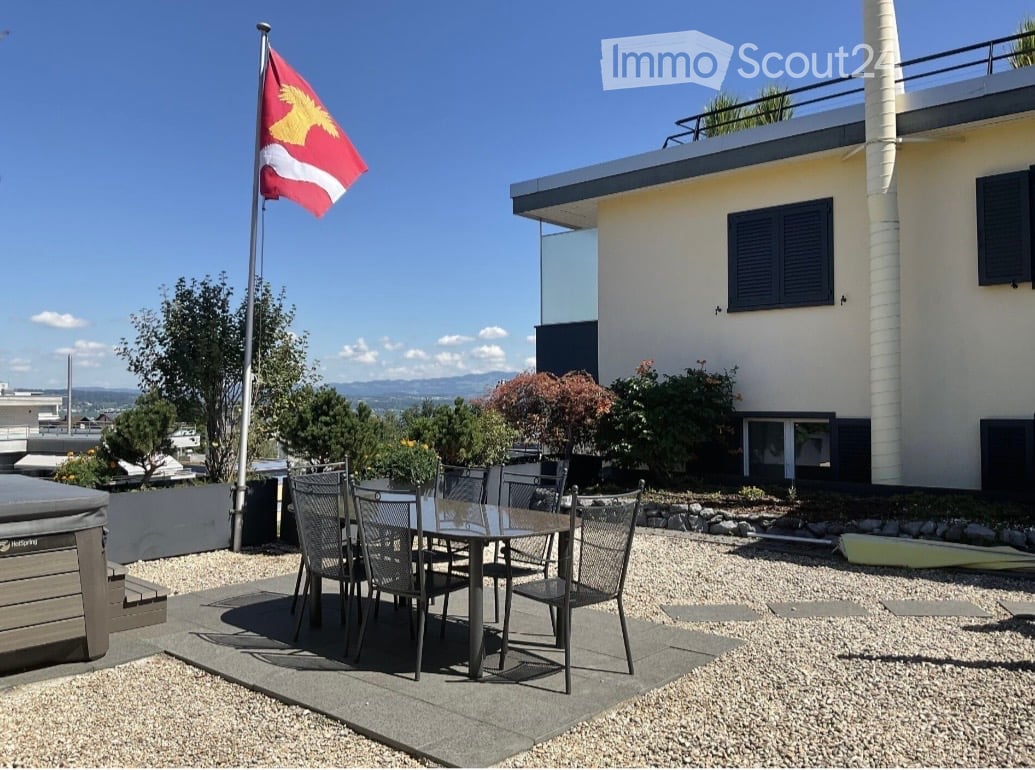 Outdoor patio area with a dining table and chairs, surrounded by gravel and small trees, with a modern house in the background and a flagpole with a flag.