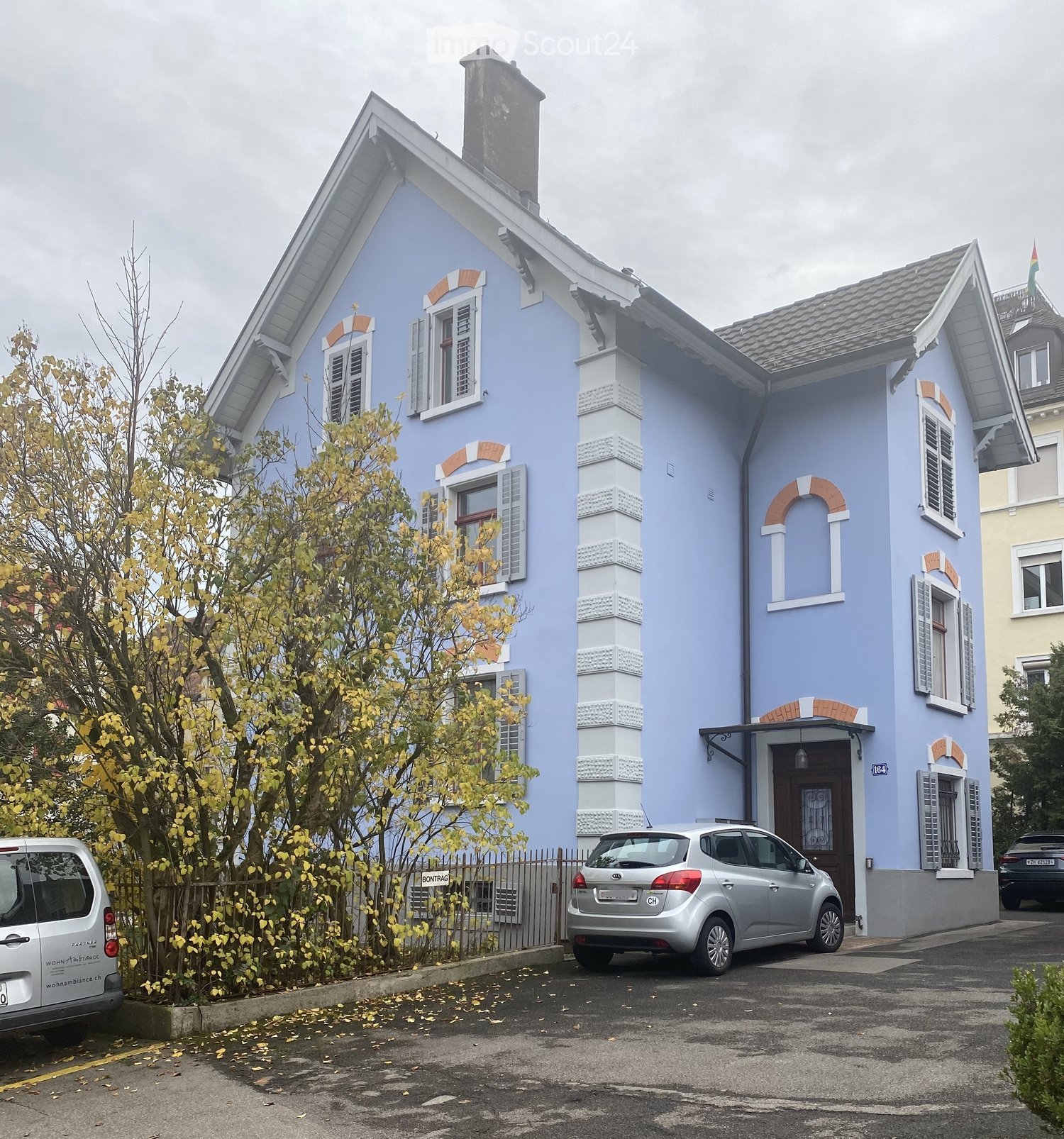 Two-story blue house with white trim, multiple windows, and a chimney. A silver car is parked in front, and a tree is visible to the left.