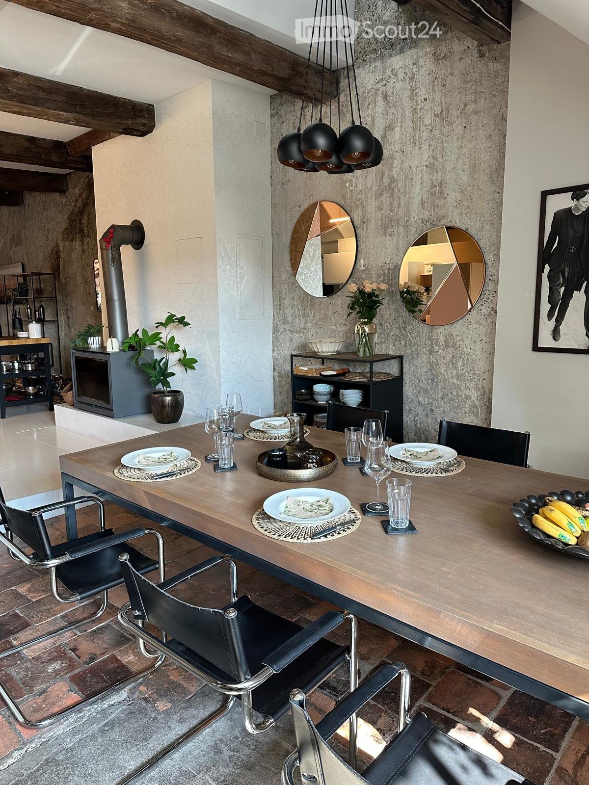 Modern dining area with a long wooden table, black leather chairs, decorative mirrors, a vase with flowers, and a fireplace in the background.