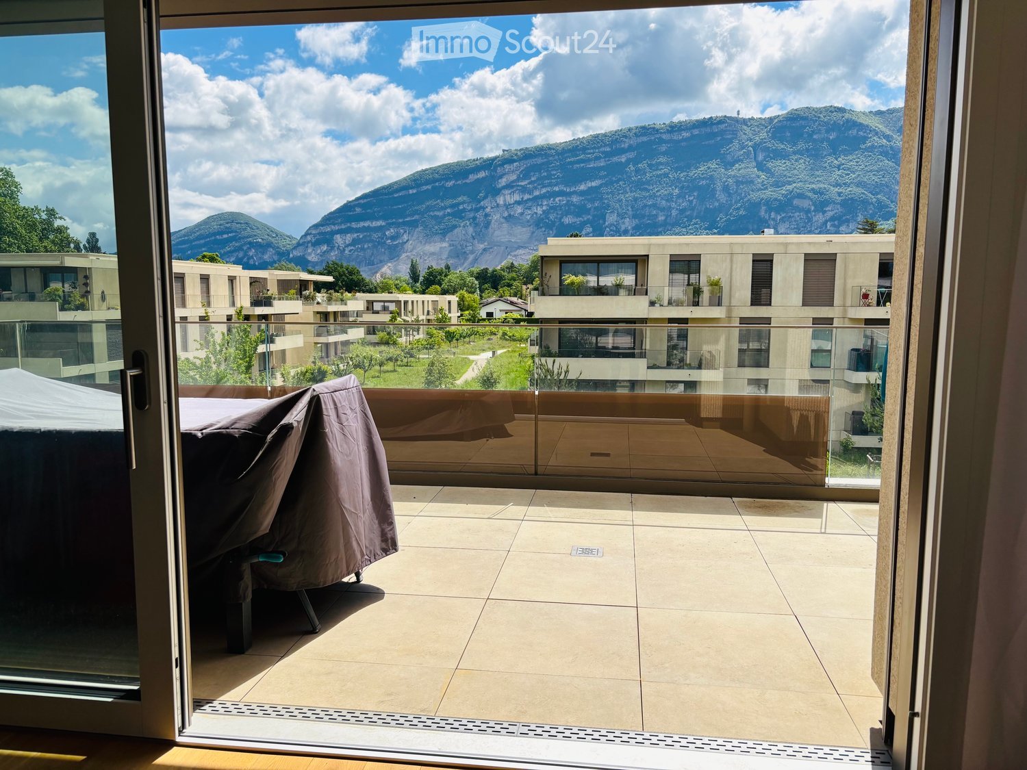 Balcony with tiled floor, glass railing, covered chair, and scenic mountain view.