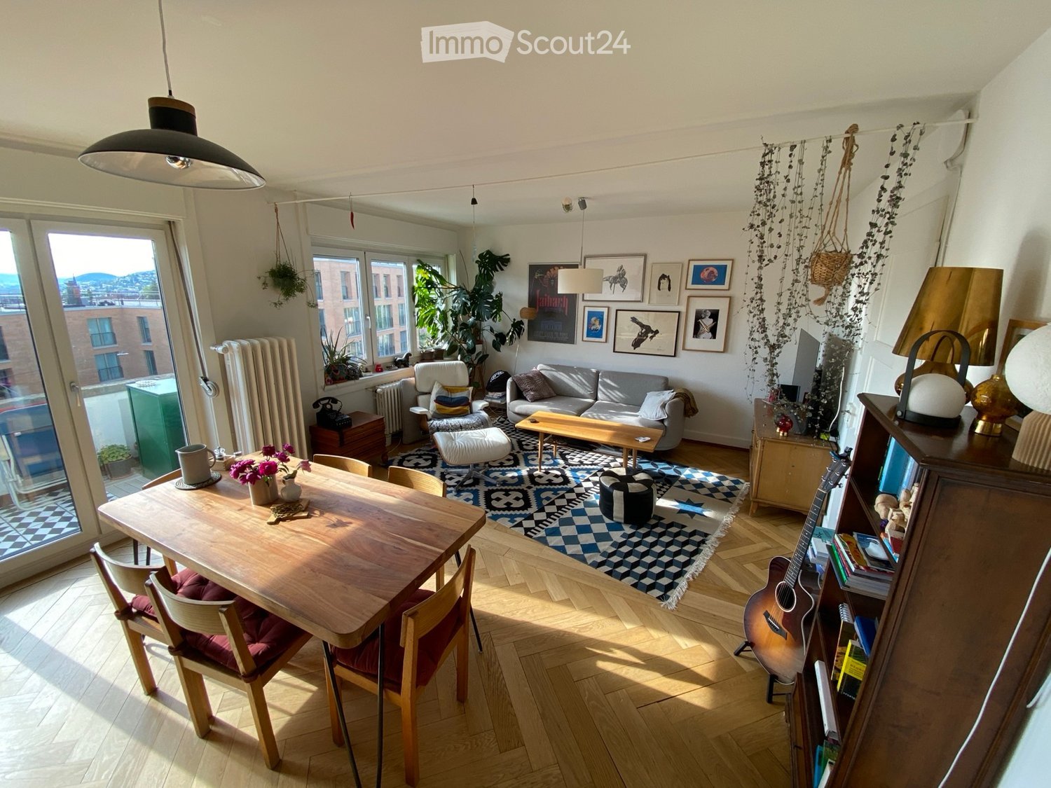 Bright living room with wooden flooring, large windows, a sofa, coffee table, dining table with chairs, guitar, bookshelf, and decorative items.