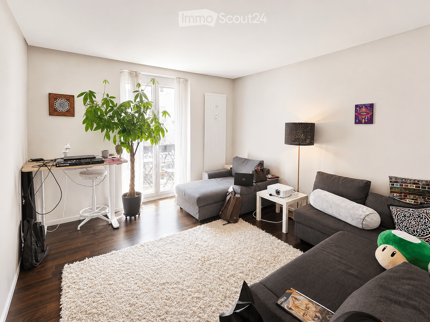 Modern living room with grey sectional sofa, white shag rug, coffee table, floor lamp, potted plant, and a small desk with a laptop and stool.