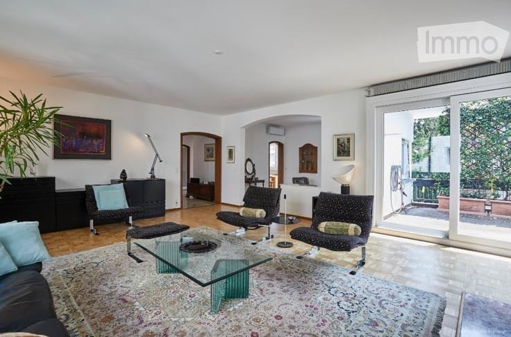 Modern living room with large windows and sliding glass doors leading to a balcony. Features a dark-colored sofa, glass coffee table, armchairs, and decorative items.