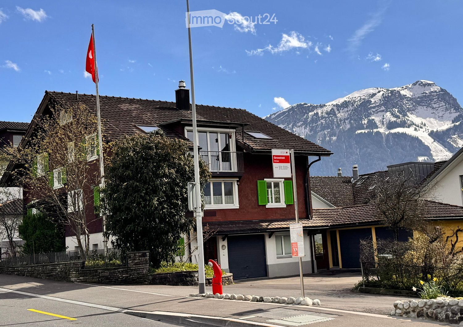 A two-story house with a red exterior, green shutters, and a balcony, situated in a picturesque village with snow-capped mountains in the background.