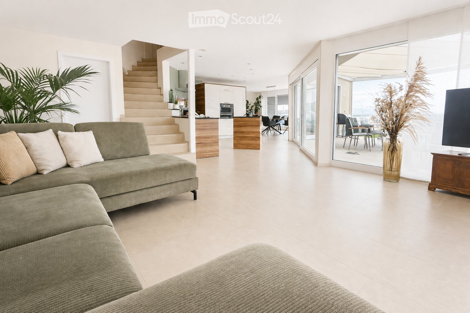 Modern living room with a green sectional sofa, ottoman, dining area with chairs and table, large glass doors leading to a balcony, and decorative vase with dried plants.