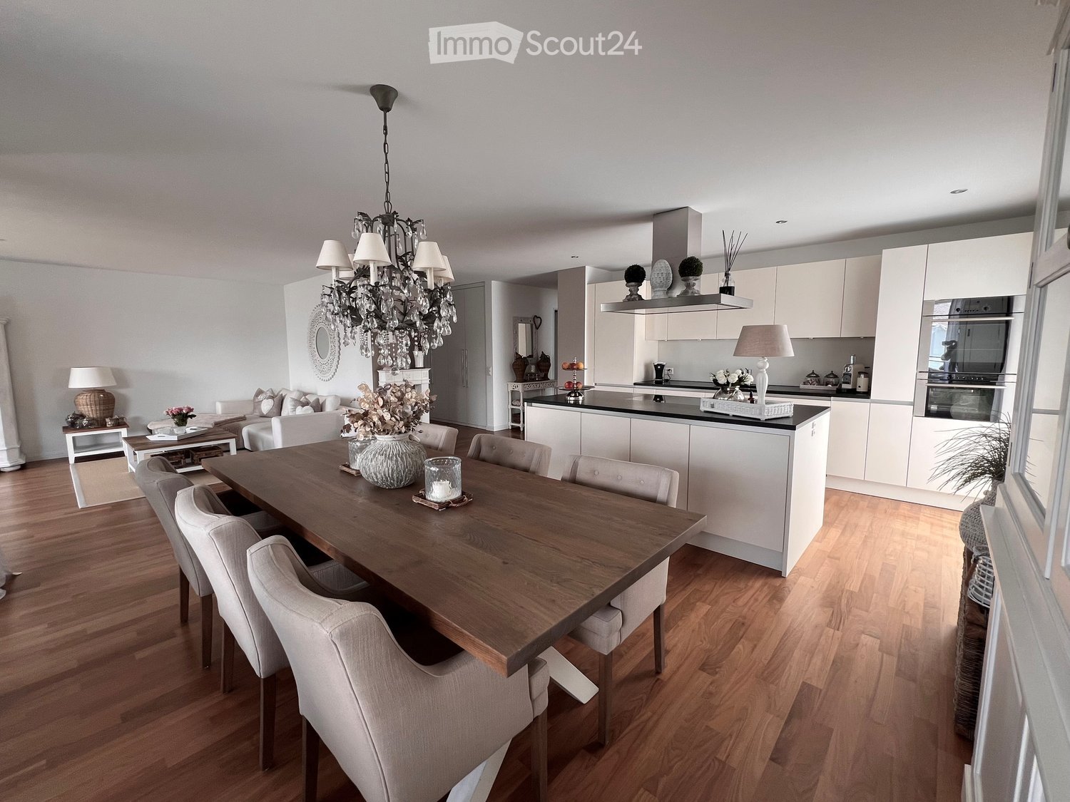 Open-plan living and dining area with a wooden dining table, six chairs, a sofa, coffee table, chandelier, and a modern kitchen with white cabinets and stainless steel appliances.