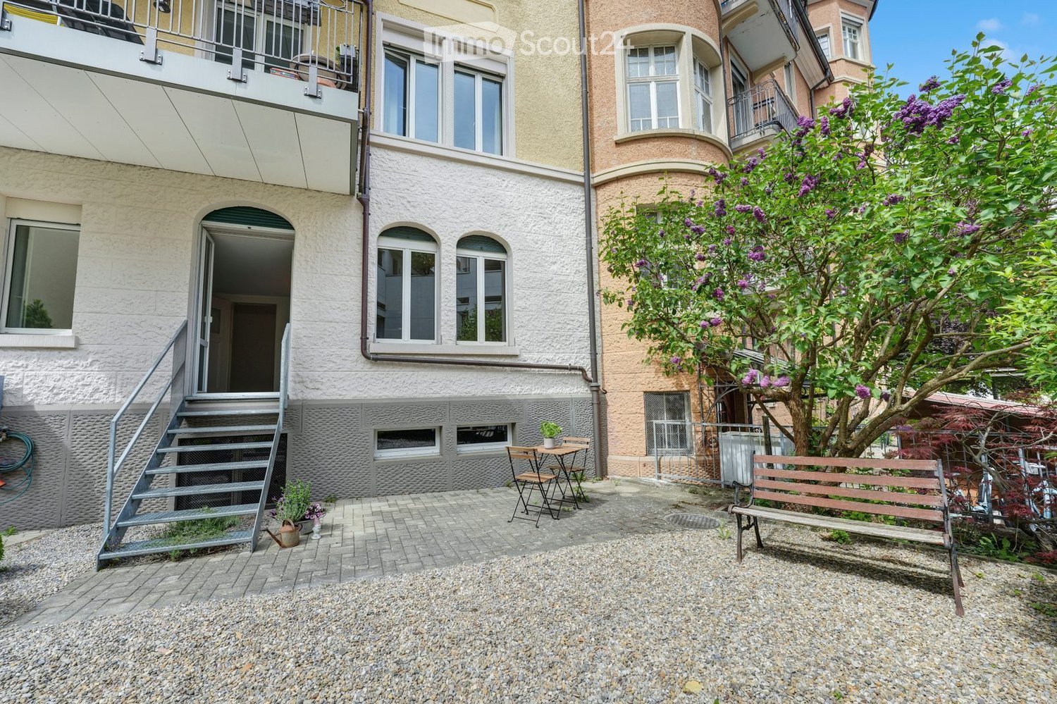 Courtyard with gravel ground, bench, small table and chairs, potted plants, and a staircase leading to a building entrance.