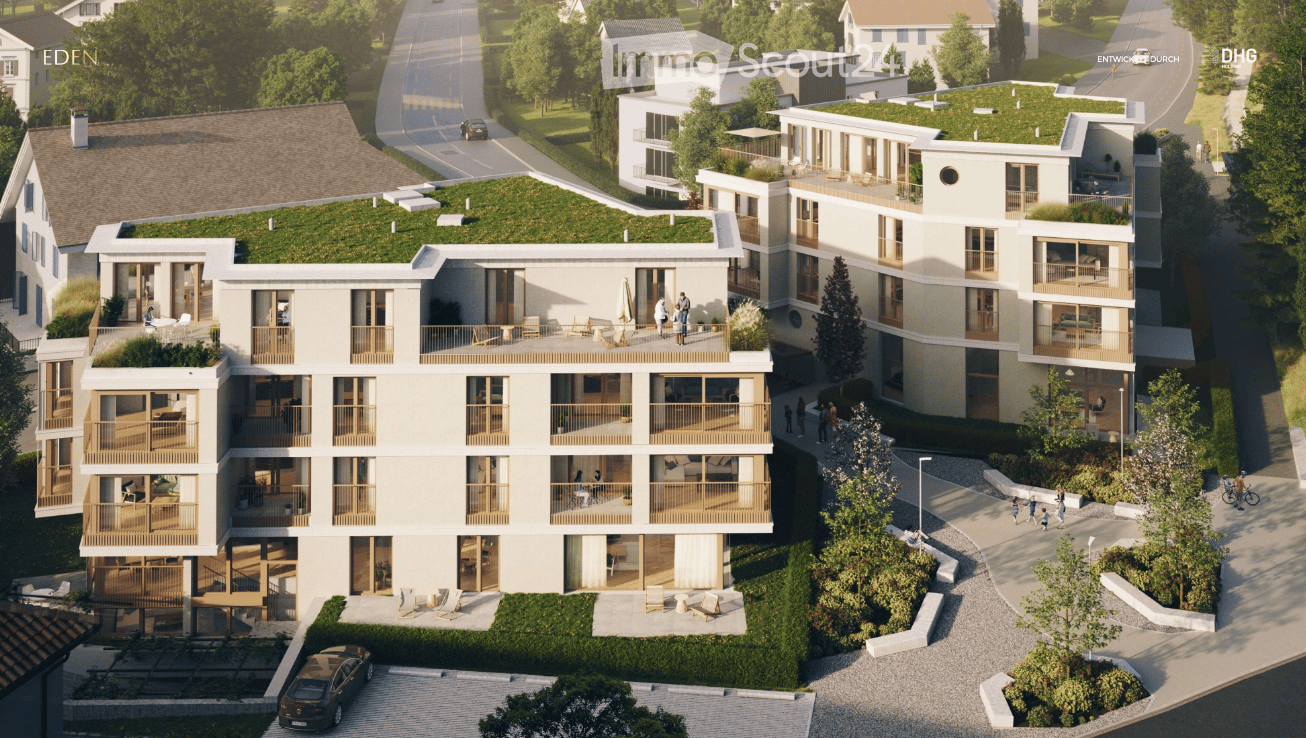 A modern multi-story residential building with balconies, green rooftop, and surrounding greenery. The building features large windows, wooden railings, and a paved parking area.