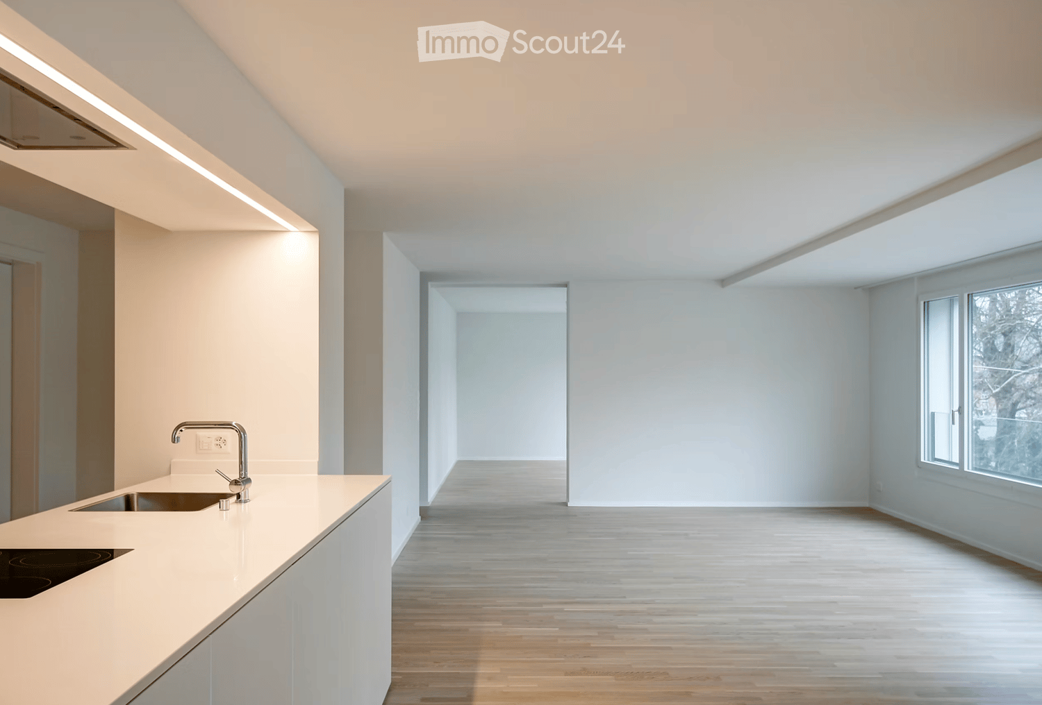 Modern, minimalist kitchen with white countertops, a built-in sink, and large windows allowing natural light. The space is empty, with no visible furniture or appliances beyond the sink and countertop.