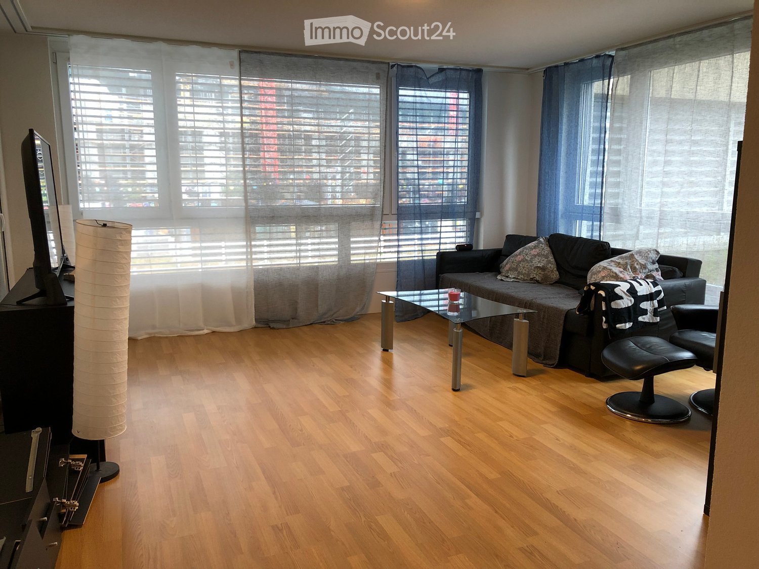 Living room with wooden flooring, large windows with blinds, a sofa with pillows, a glass coffee table, a floor lamp, and a television on a stand.