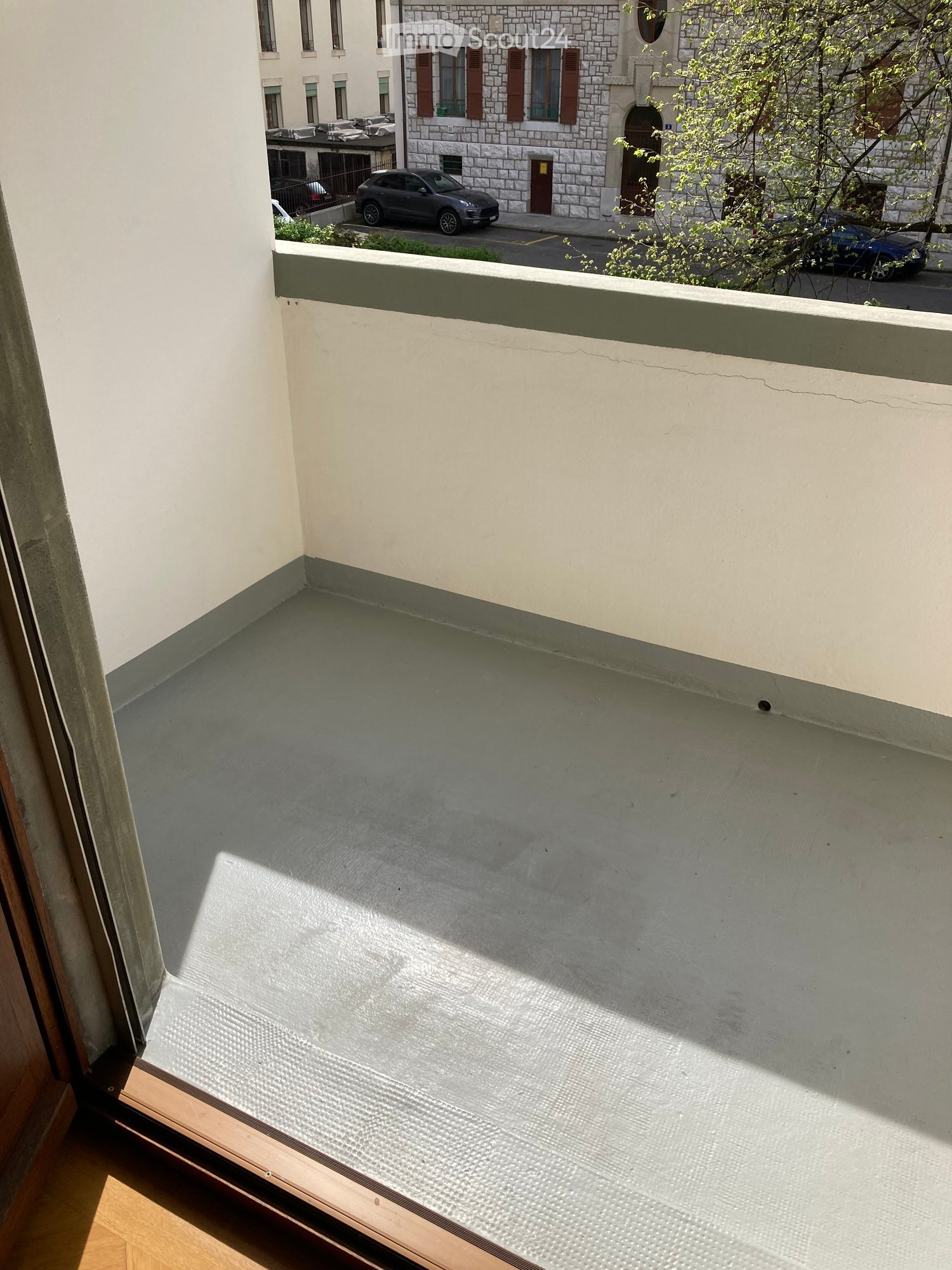 Balcony with concrete floor, white walls, and a view of a street with parked cars and a building with stone facade and shutters.