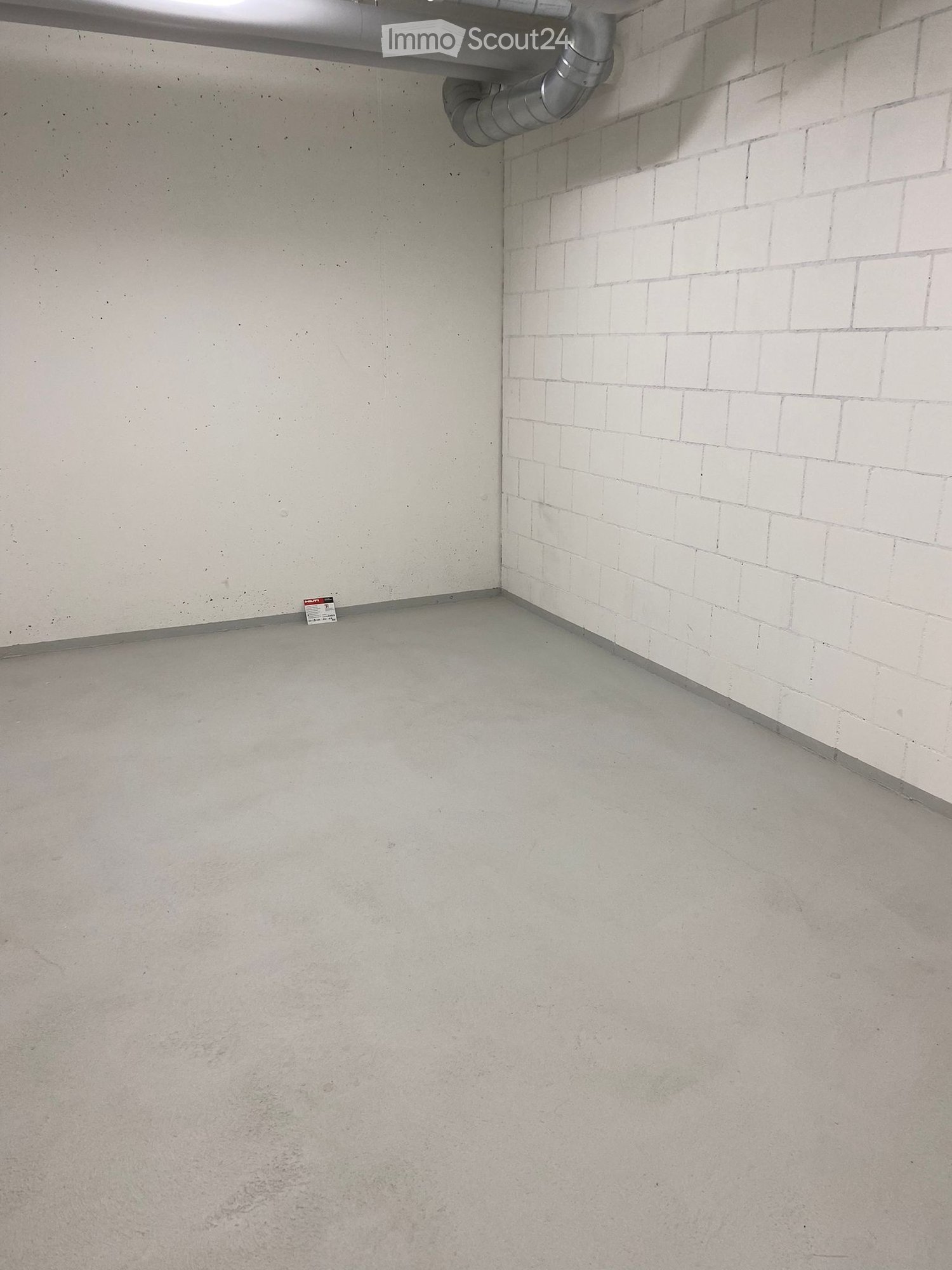 Empty room with concrete floor and white tiled wall, featuring a metal pipe and a small sign on the floor.