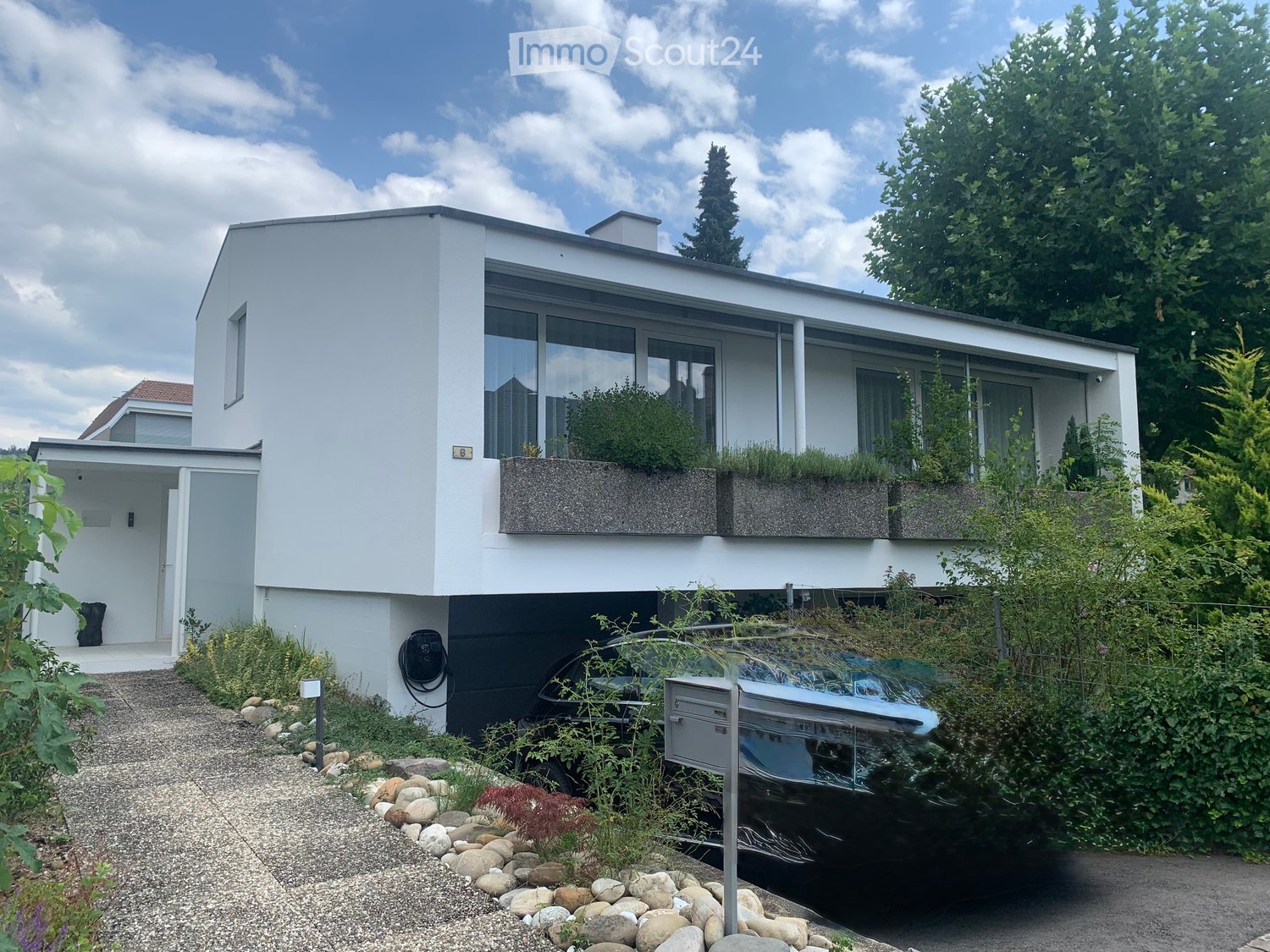Modern two-story house with white exterior, large windows, and a flat roof. Features a small balcony with greenery, a driveway, and a parked car.