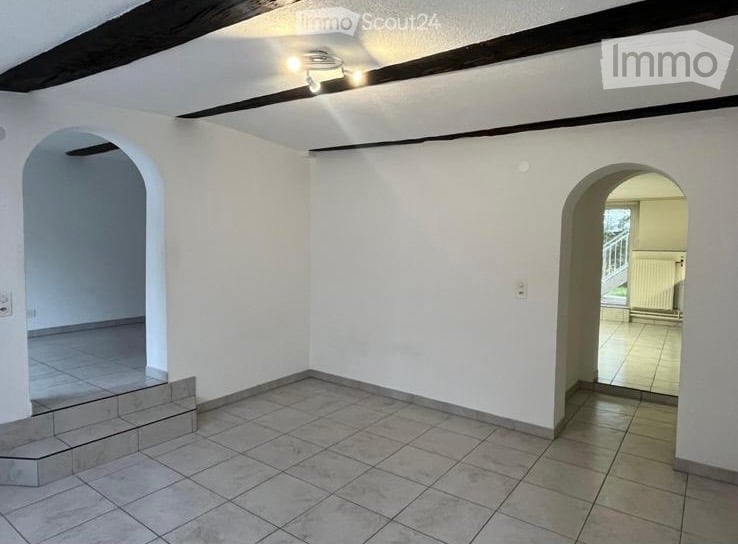 Empty room with white walls, tiled floor, and two arched doorways leading to other spaces. The room has a raised platform on one side and a ceiling with exposed wooden beams and a central light fixture.