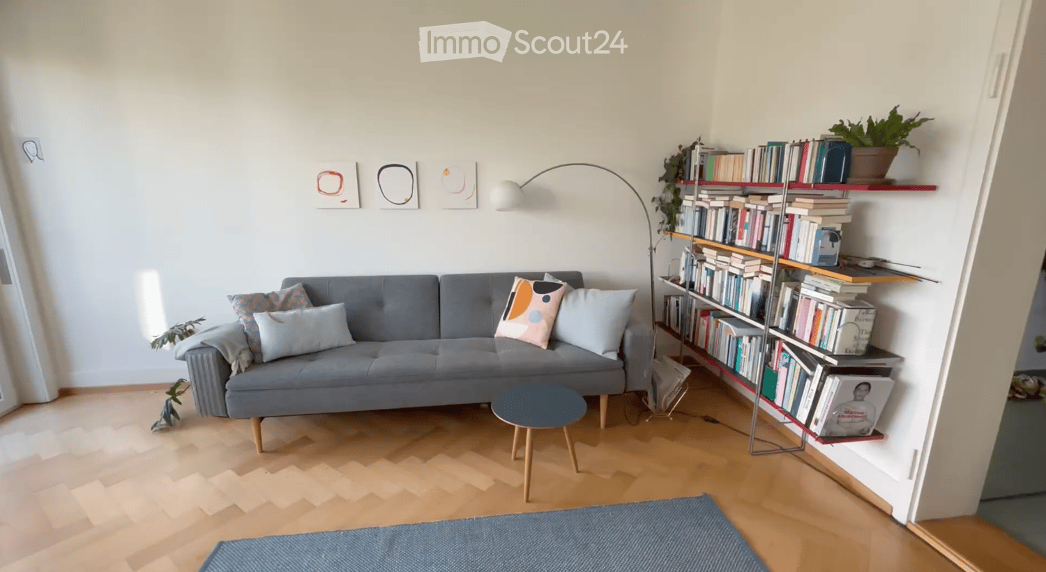 Modern living room with a grey sofa, wooden coffee table, bookshelf filled with books, floor lamp, and decorative pillows.