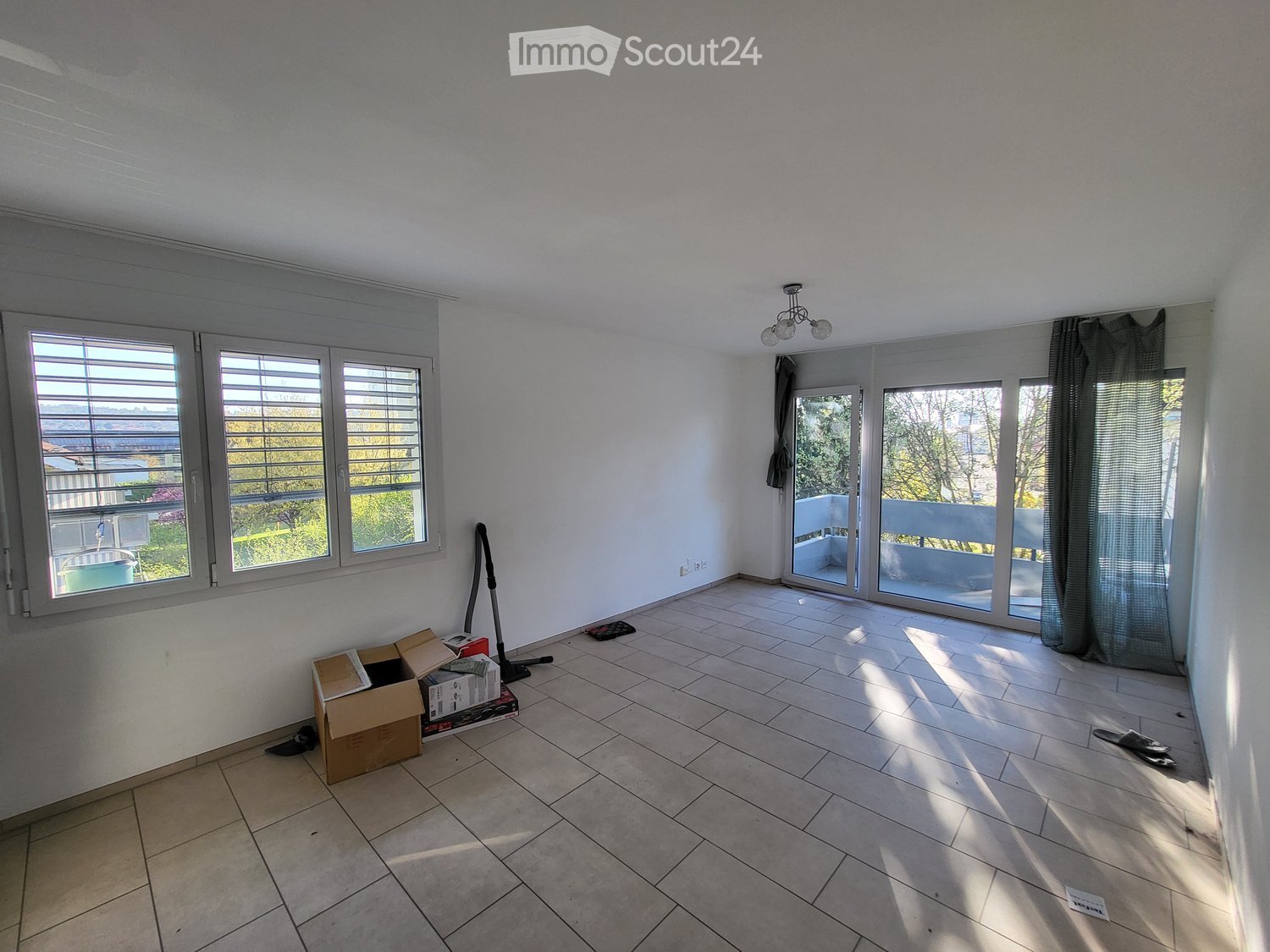 Empty room with tiled floor, large windows, sliding glass doors leading to a balcony, and some scattered items like a vacuum cleaner and boxes.