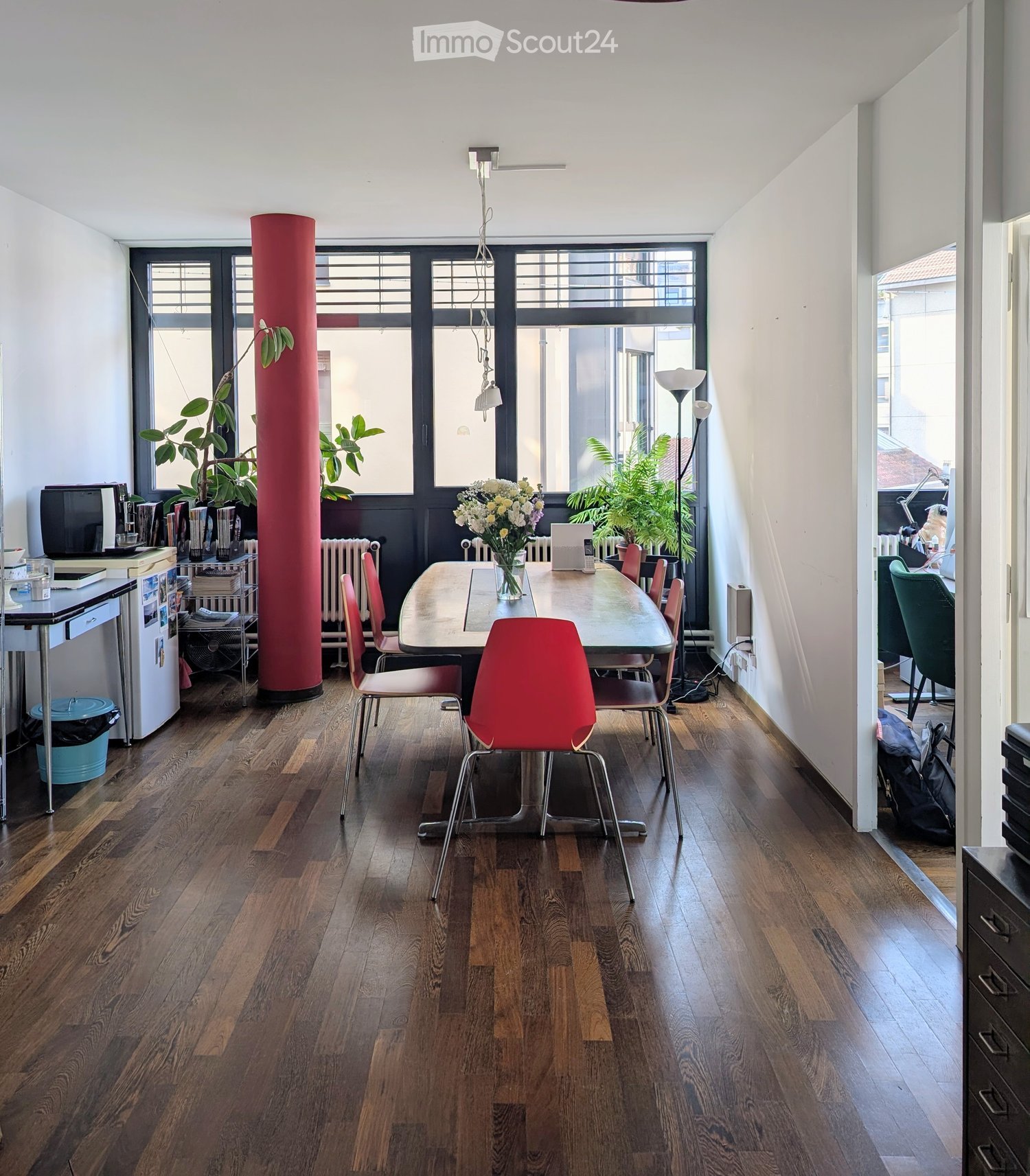 A modern, well-lit room with wooden flooring, large windows, and a central dining table surrounded by colorful chairs. There is a refrigerator, a fan, and a laptop on a side table, along with a flower vase on the dining table.