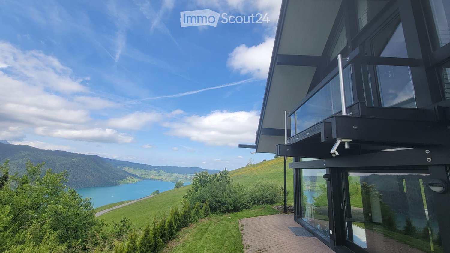 Modern two-story house with large glass windows and a balcony overlooking a scenic lake and green hills.