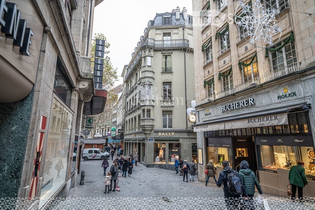 A bustling urban street lined with classic European buildings, featuring luxury shops like Bucherer and Rolex, with pedestrians walking along the cobblestone pavement.