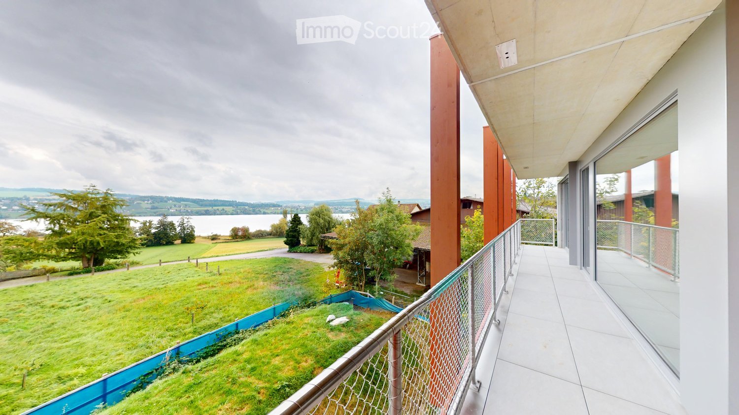 Balcony with white tiled flooring, metal railing, and a view of a green lawn, trees, and a body of water in the distance.