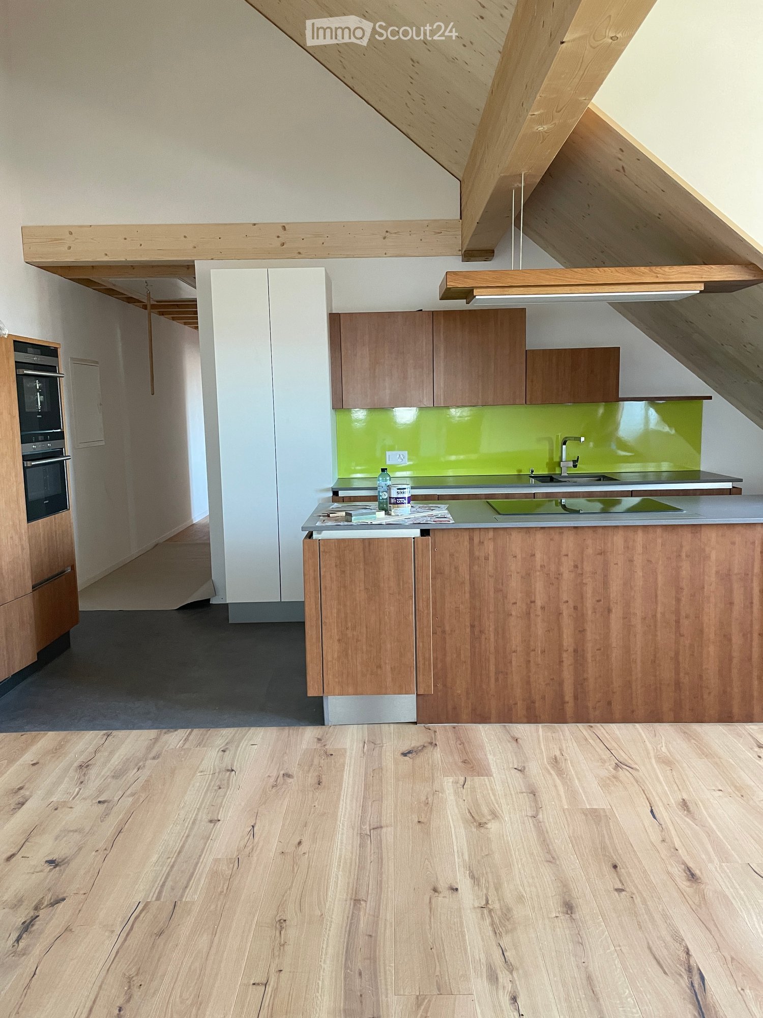 Modern kitchen with wooden cabinets, a central island, and a green backsplash. The space features a sleek design with a wooden floor and exposed wooden beams on the ceiling.