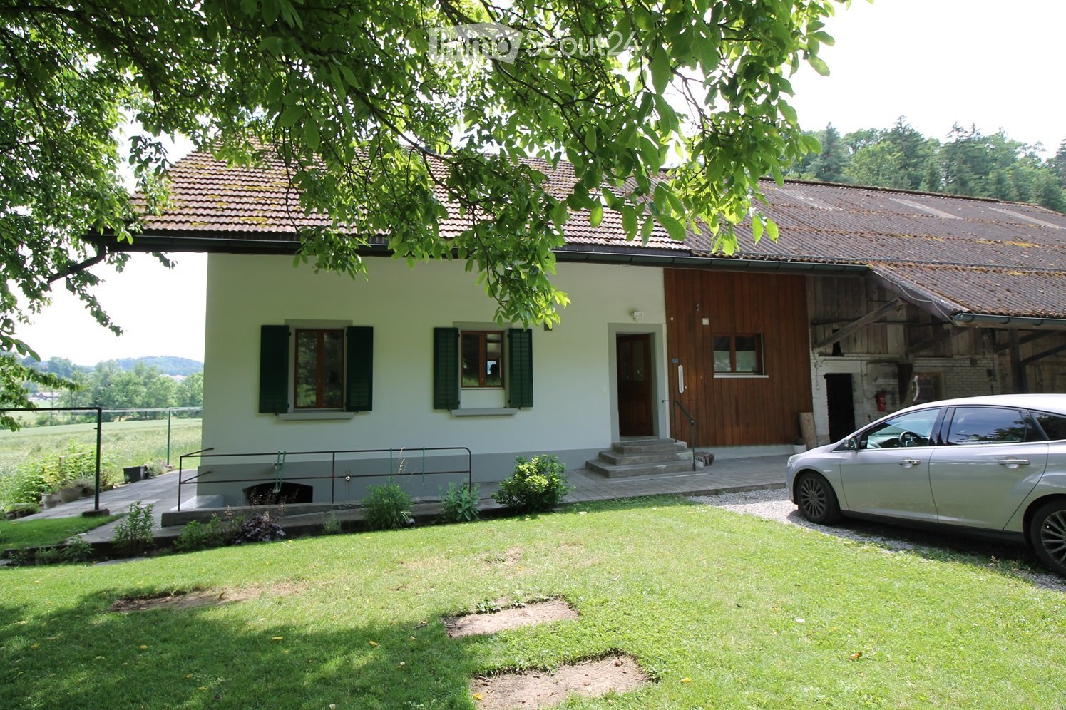 Two-story house with light green exterior, wooden shutters, and a tiled roof. A silver car is parked on a gravel driveway, and the house is surrounded by a well-maintained lawn and trees.