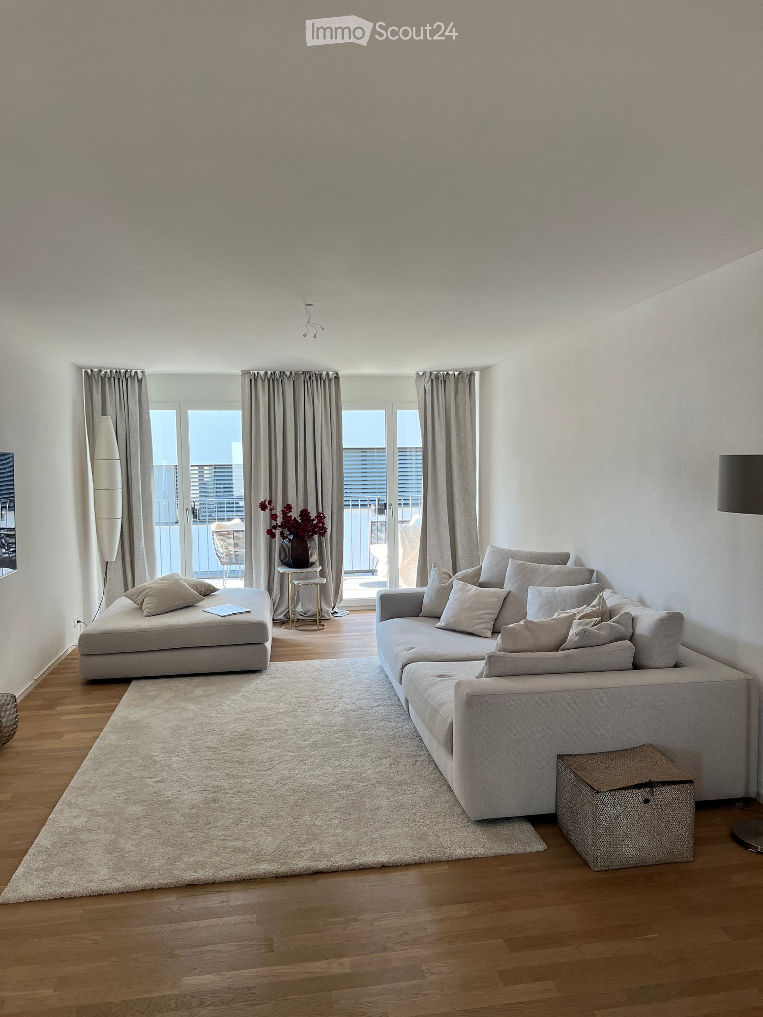 Modern living room with light grey sofa, ottoman, small round table with vase, floor lamp, and large windows leading to a balcony.