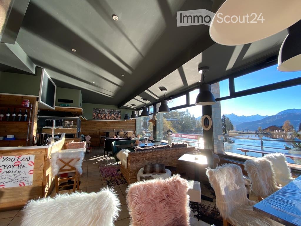 Modern lounge area with large windows offering a scenic mountain view, featuring a wicker sofa, plush chairs, a small table, and a bar area with a screen.