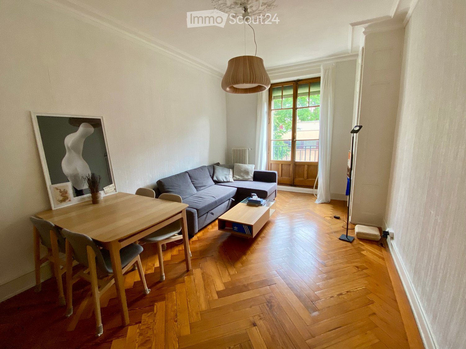 Bright living room with wooden flooring, a grey sectional sofa, a wooden coffee table, a dining table with chairs, and a large mirror on the wall.
