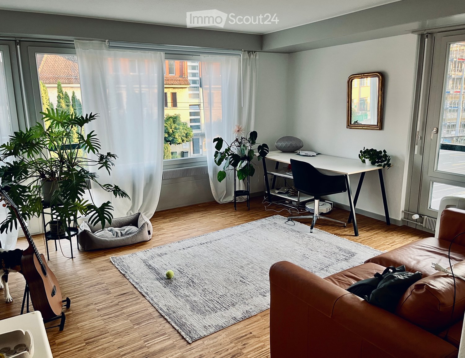 Modern living room with wooden flooring, large windows, a brown leather sofa, a guitar, a pet bed, a desk with a chair, and several plants.