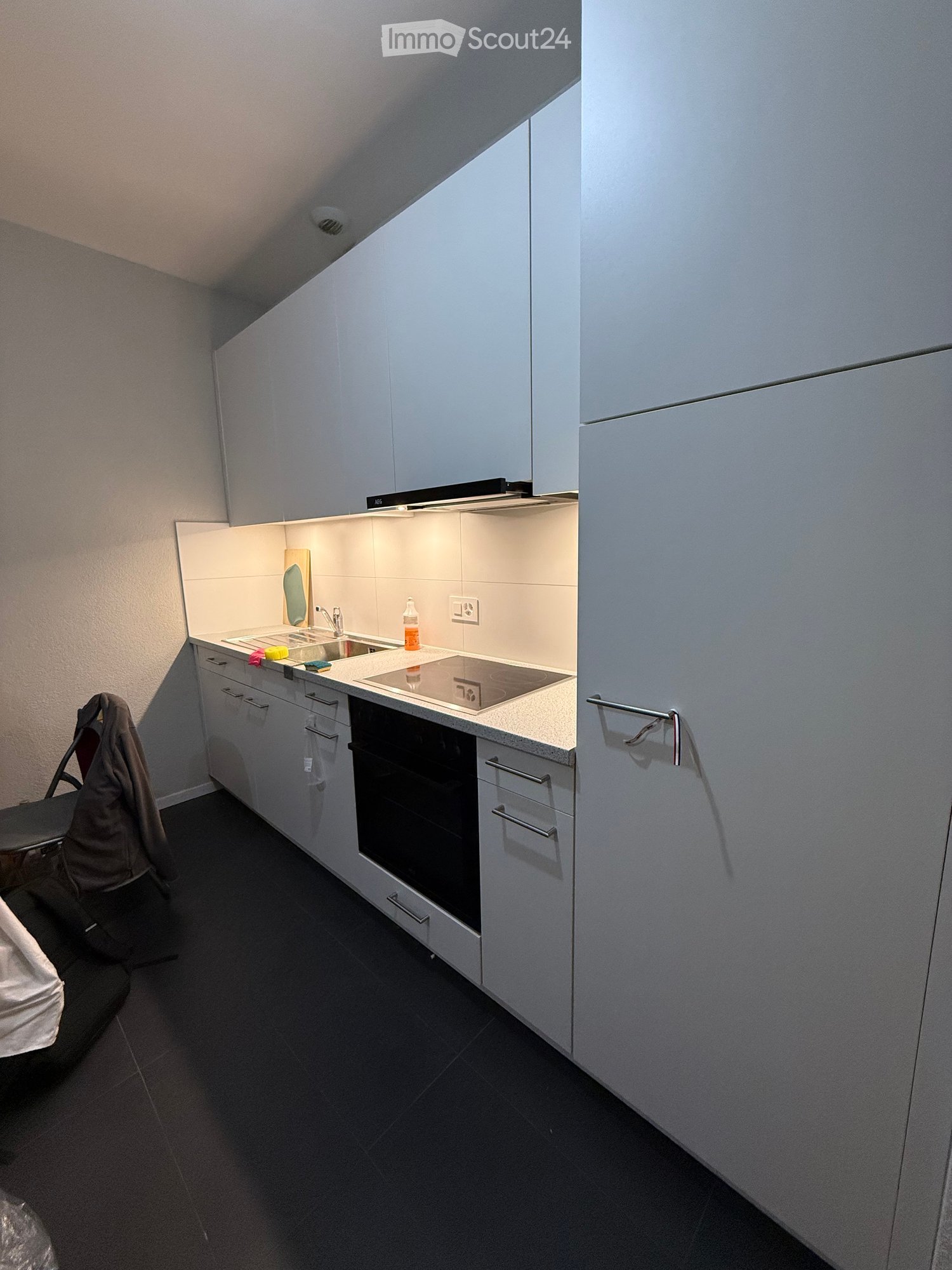Modern kitchen with white cabinets, dark countertops, built-in oven, and sink area. Includes a bottle on the counter and a jacket draped over a chair.