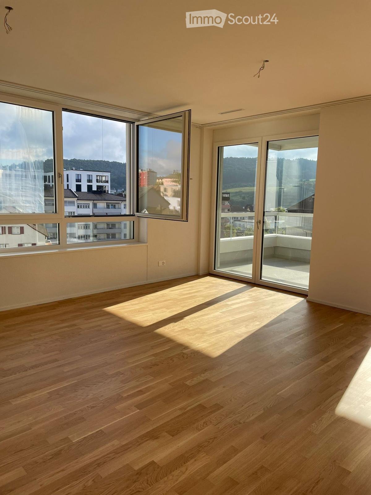 Empty room with wooden flooring, large windows, and sliding glass doors leading to a balcony.