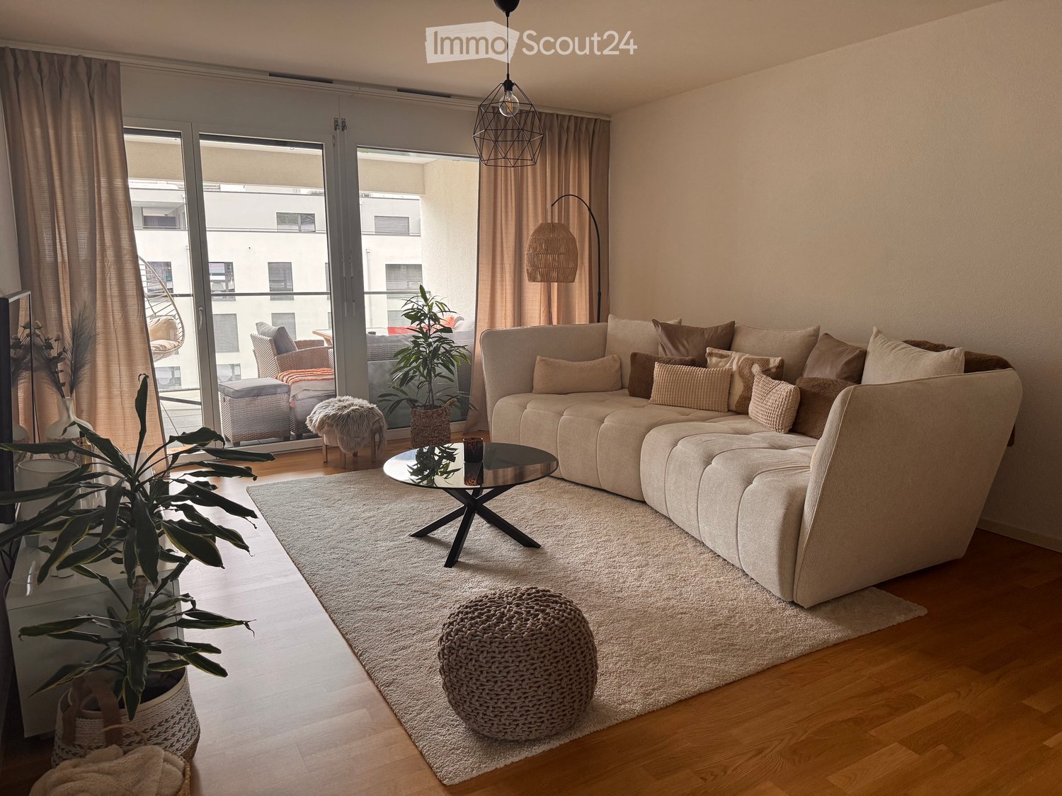 Modern living room with a large sectional sofa, coffee table, round stool, potted plants, and a view of a balcony area.