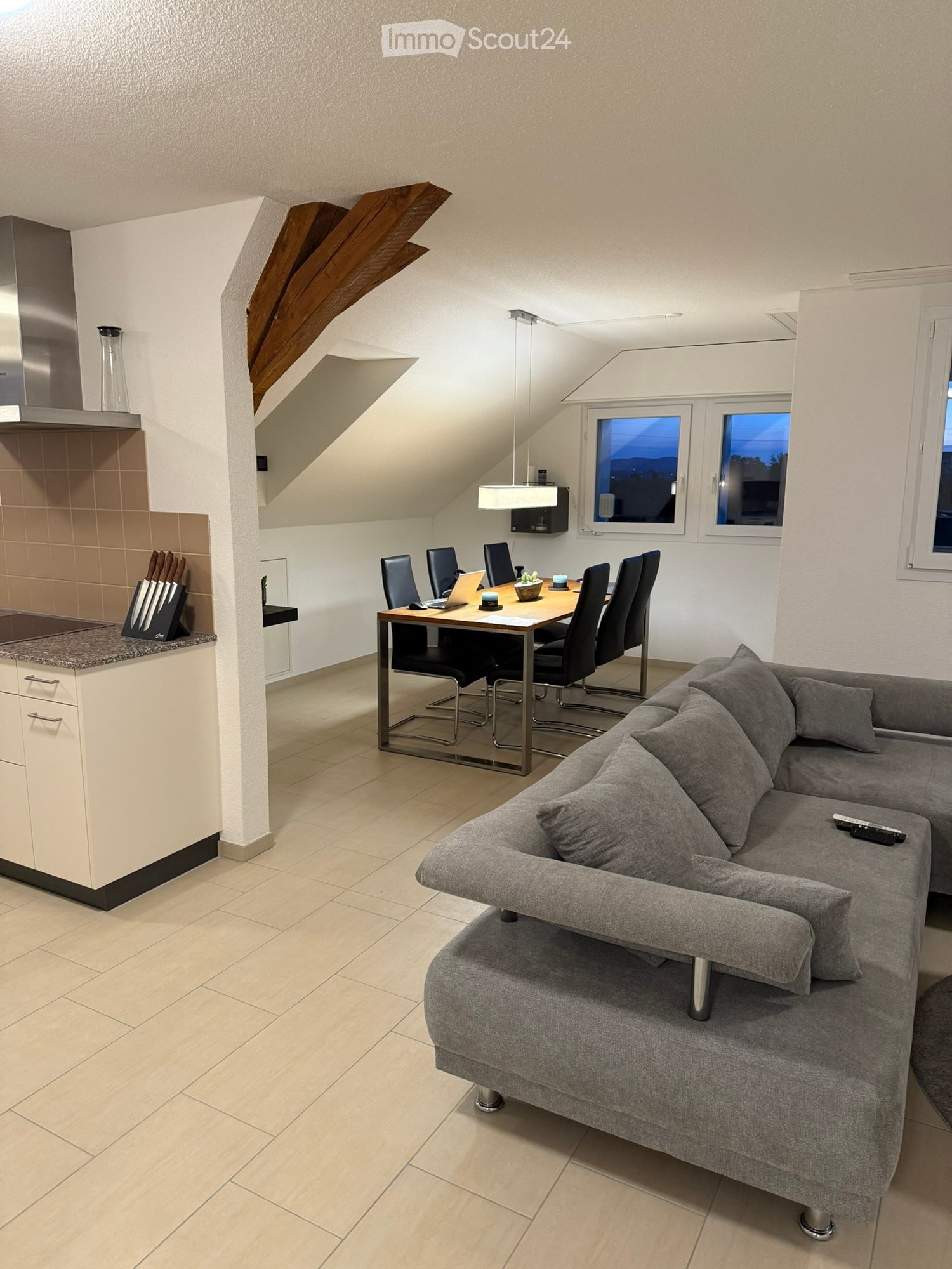 Modern, open-concept living and dining area with a grey sectional sofa, a dining table with six chairs, a laptop, and a small kitchen area featuring a countertop, cabinets, and a knife block. The space has large windows and a slanted ceiling with exposed