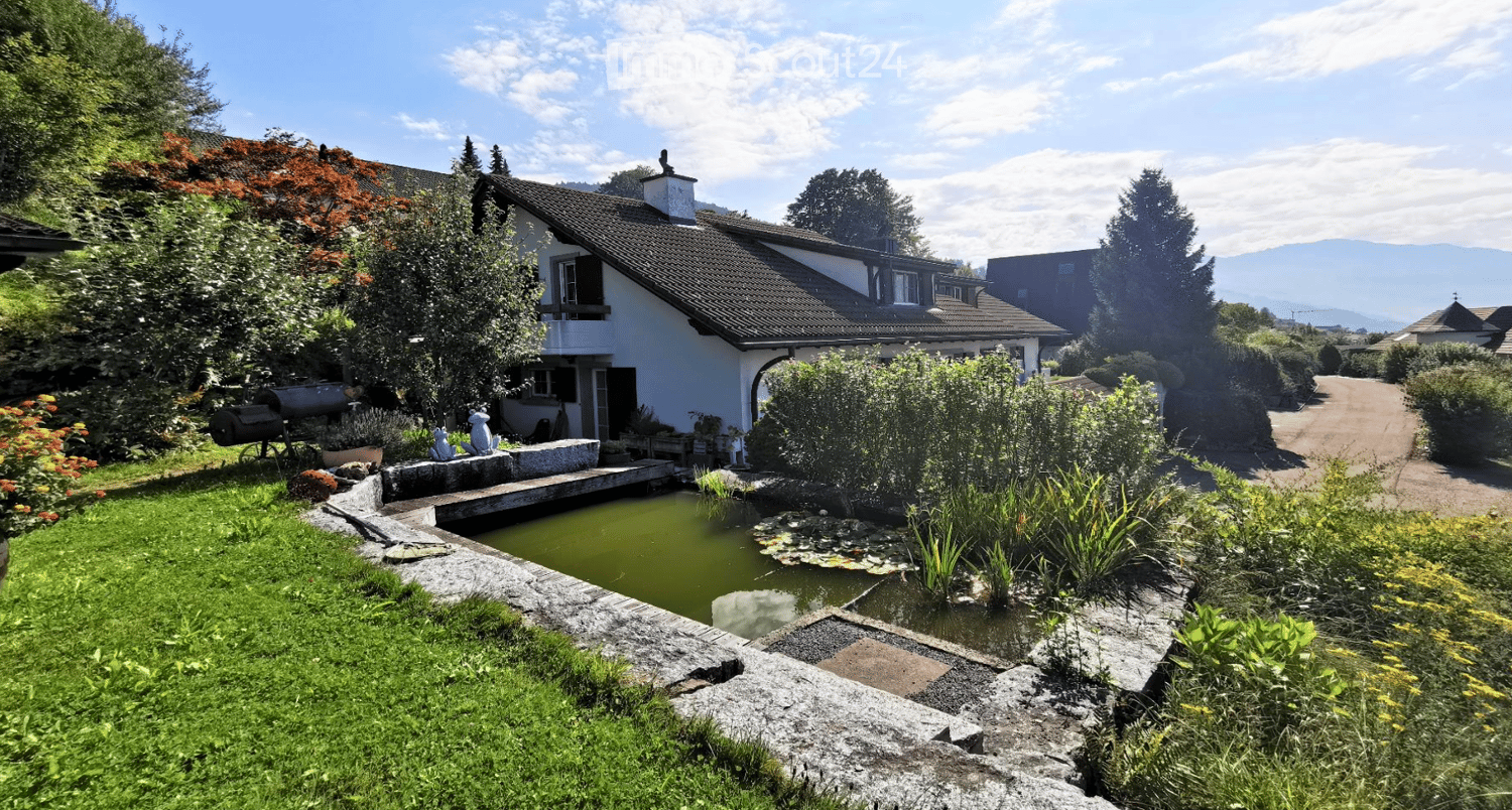 A serene garden area with a small pond, surrounded by lush greenery, a stone pathway, and a charming white house with a sloped roof in the background.