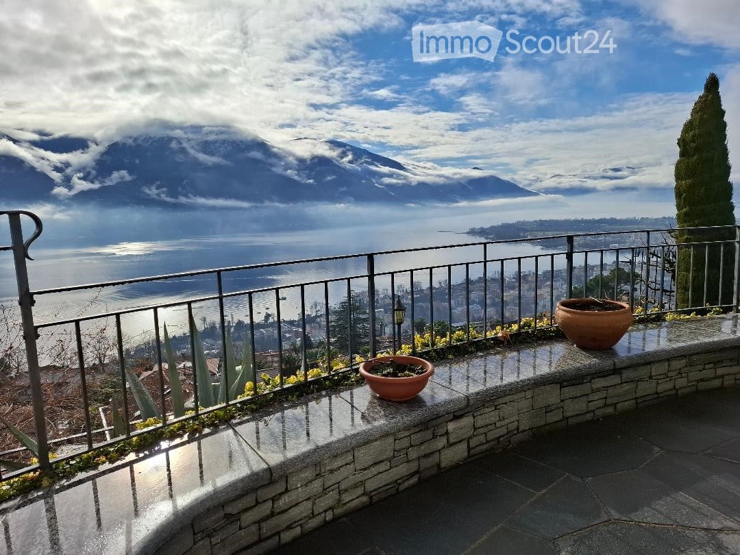 Terrace with black railings, concrete walkway, and potted plants, overlooking a sea and mountains