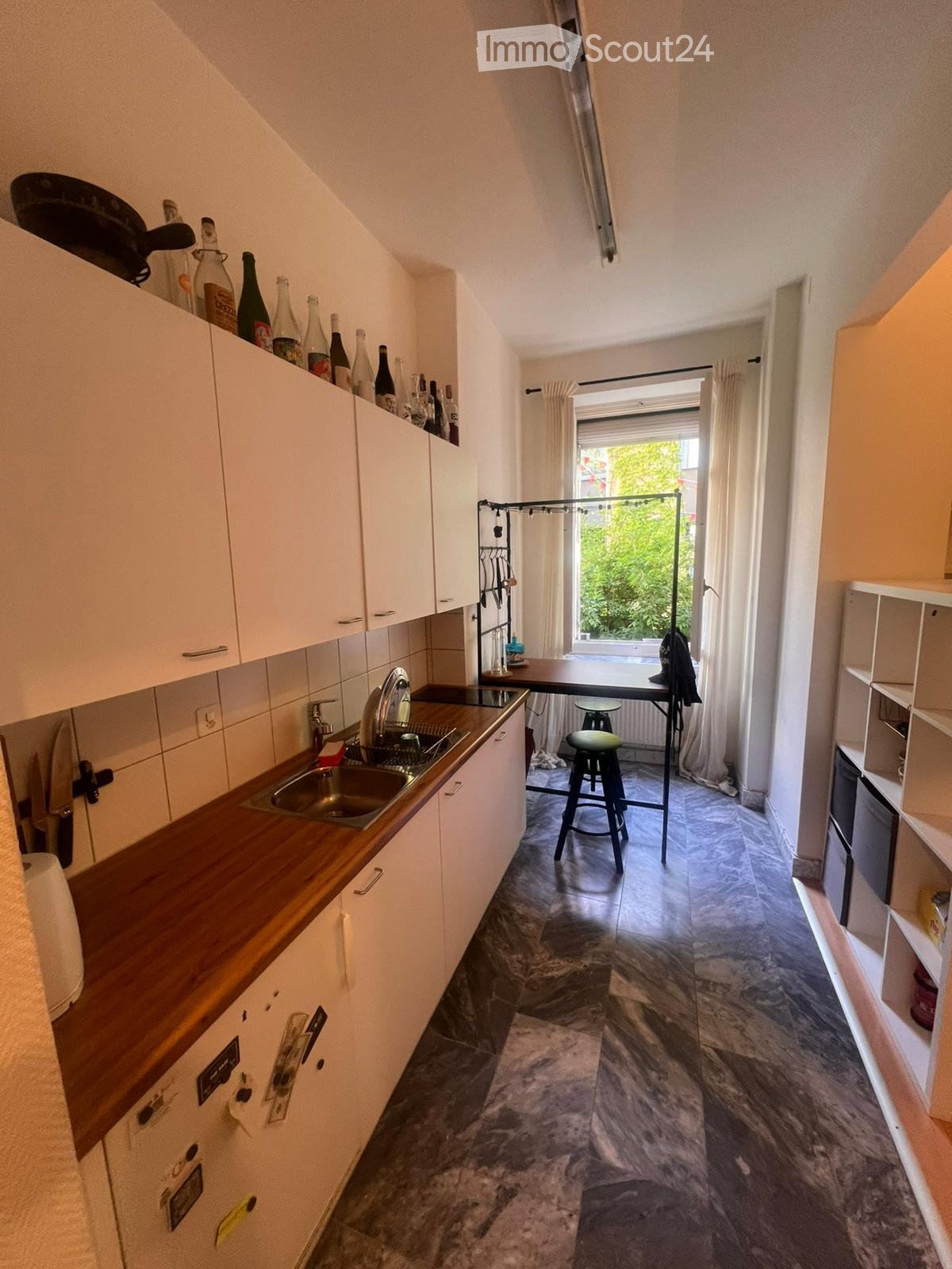 The kitchen features white cabinets, a wooden countertop, a sink with a faucet, and a small table with a stool. Bottles are placed on top of the cabinets, and a curtain covers the window.