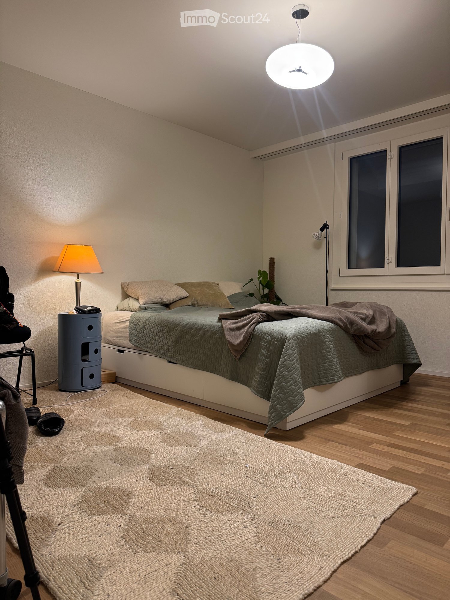 Cozy bedroom with a double bed, beige bedding, a bedside table with a lamp, a large round ceiling light, a woven rug, and a small plant near the window.