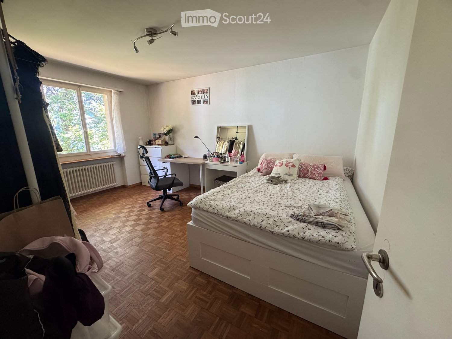 Cozy bedroom with wooden flooring, a single bed with pillows and a blanket, a desk with a chair, a mirror, and a radiator under the window.