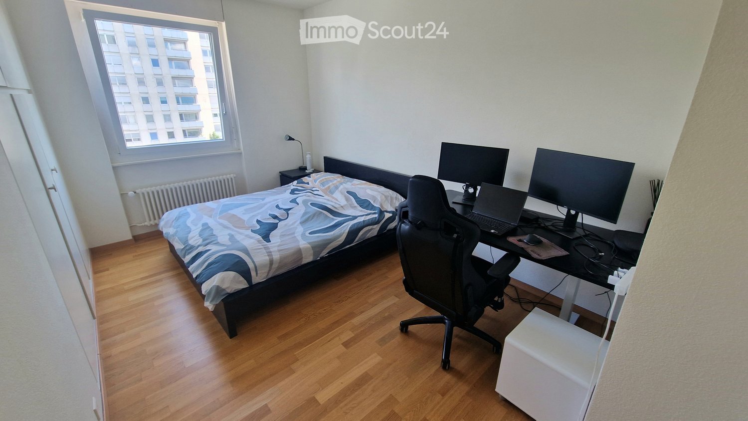 A modern, furnished room with a bed, desk, laptop, and two large screens. The room features wooden flooring and large windows with a city view.