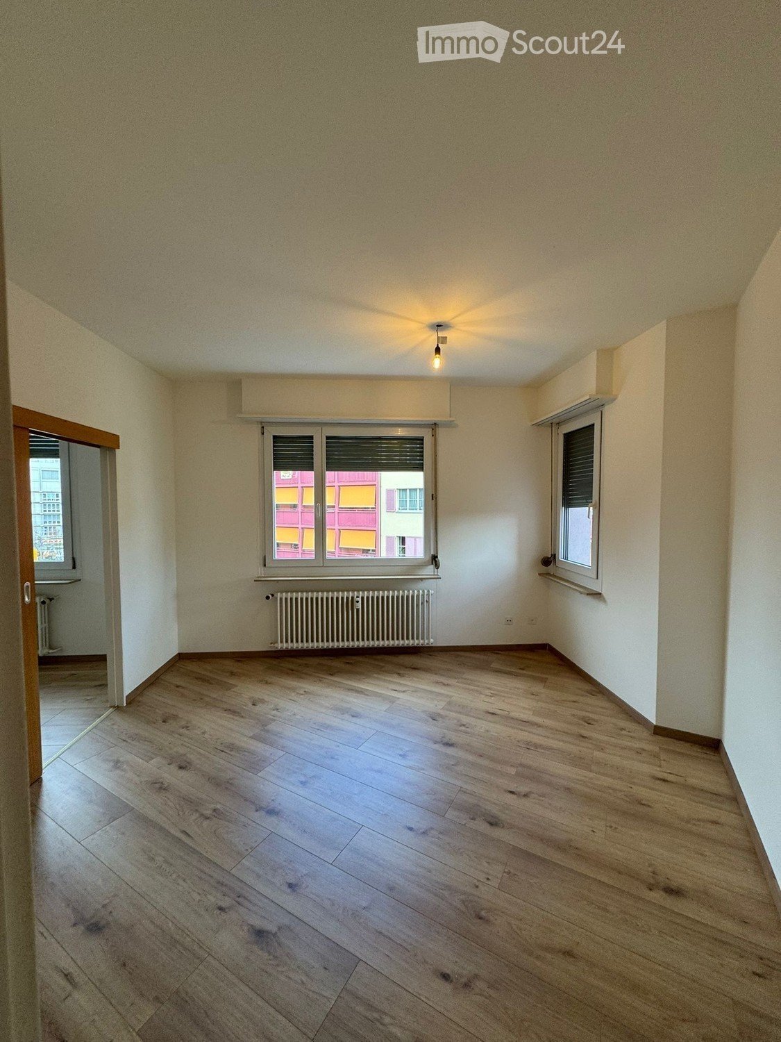 Empty room with wooden flooring, white walls, and large windows with blinds. A radiator is visible below one window, and a single ceiling light fixture is present.
